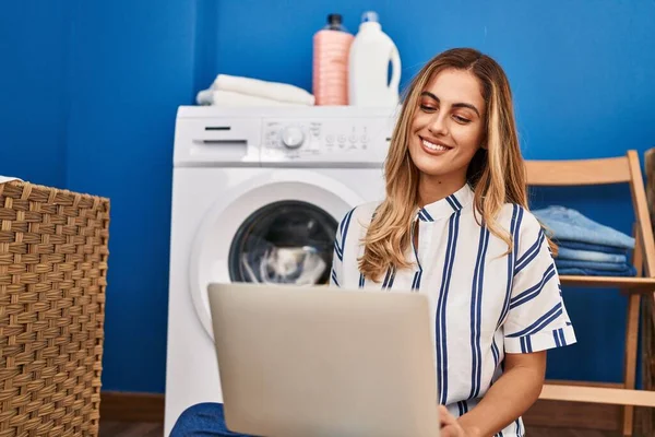 年轻的金发女人在洗衣房用手提电脑等洗衣机 — 图库照片