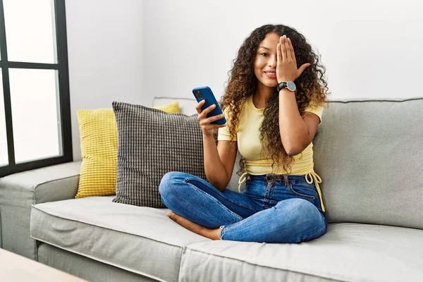 自宅のソファに座っている美しいヒスパニック系の女性は 手で一つの目を覆うスマートフォンを使用して 顔に自信を持って笑顔と驚きの感情 — ストック写真