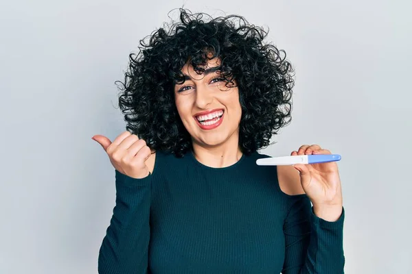 Young middle east woman holding pregnancy test result pointing thumb up to the side smiling happy with open mouth