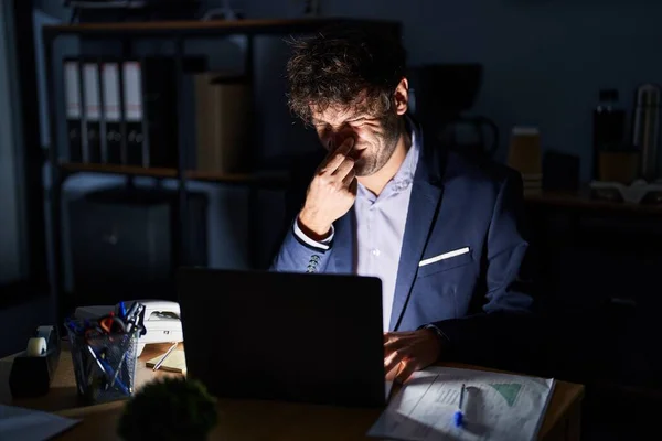 Hispanic young man working at the office at night smelling something stinky and disgusting, intolerable smell, holding breath with fingers on nose. bad smell