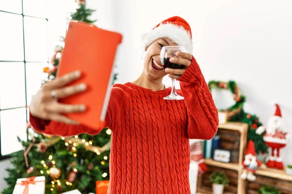 Young hispanic woman having video call toasting with wine standing by christmas tree at home