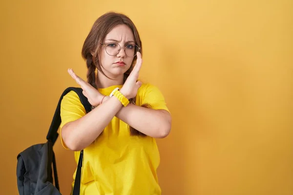 Young caucasian woman wearing student backpack over yellow background rejection expression crossing arms doing negative sign, angry face