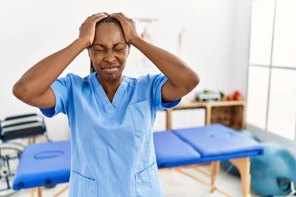 Black woman with braids working at pain recovery clinic suffering from headache desperate and stressed because pain and migraine. hands on head.