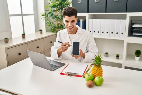 Joven Hispano Vistiendo Uniforme Dietista Mostrando Teléfono Inteligente Pantalla Clínica —  Fotos de Stock