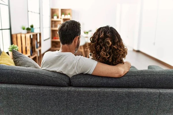 Middle age hispanic couple on back view hugging and sitting on the sofa at home.