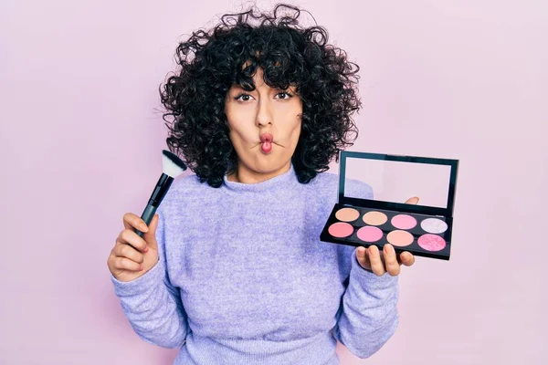 Young middle east woman holding makeup brush and blush making fish face with mouth and squinting eyes, crazy and comical.