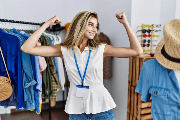 Young blonde woman working as manager at retail boutique showing arms muscles smiling proud. fitness concept.
