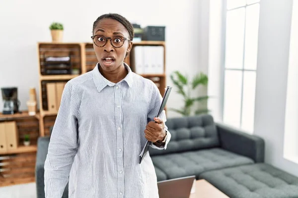 African woman working at psychology clinic in shock face, looking skeptical and sarcastic, surprised with open mouth