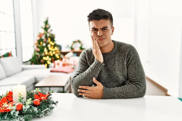 Mladý Hispánec Sedící Stole Vánočního Stromku Dotýkající Rukou Úst Bolestivým — Stock fotografie