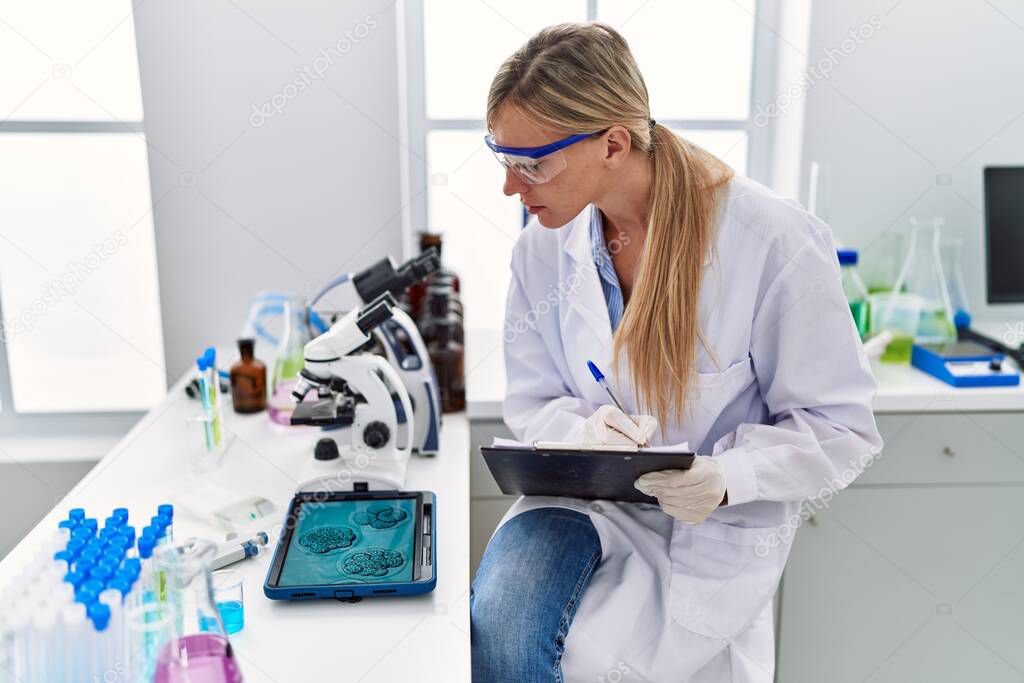 Young blonde woman wearing scientist uniform writing on clipboard looking embryion images at laboratory