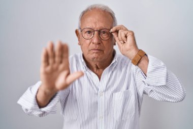 Senior man with grey hair holding glasses with hand with open hand doing stop sign with serious and confident expression, defense gesture 