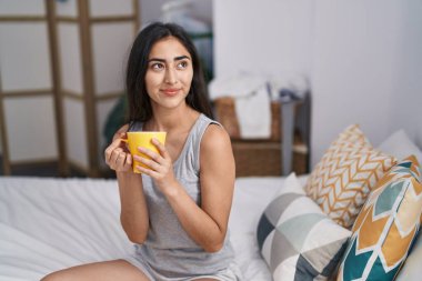 Young hispanic girl drinking cup of coffee sitting on bed at bedroom