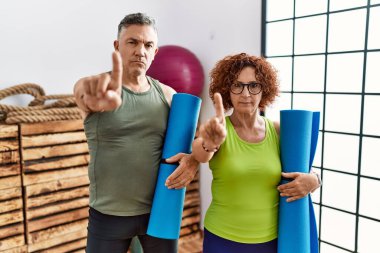 Orta yaşlı çift, yoga minderi tutarken parmağını kaldırıyor ve kızgın bir ifade takınıyor. 