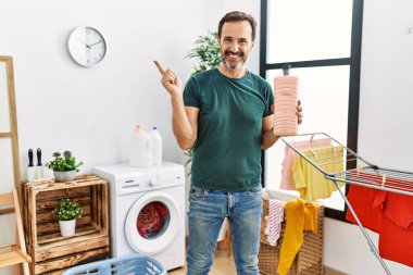 Middle age man with beard doing laundry holding detergent bottle smiling happy pointing with hand and finger to the side 