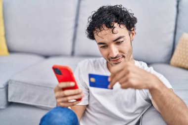 Young hispanic man using smartphone and credit card sitting on floor at home
