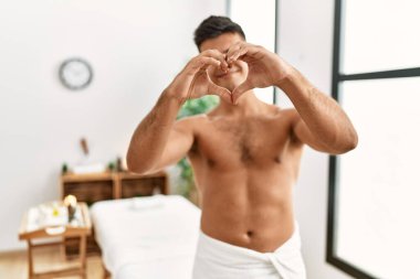 İspanyol asıllı genç adam spa merkezinde üstsüz duruyor. Sevgiyle gülümsüyor. Elleriyle kalp sembolü yapıyor. romantik kavram. 