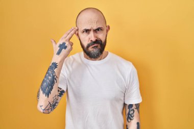 Young hispanic man with tattoos standing over yellow background shooting and killing oneself pointing hand and fingers to head like gun, suicide gesture. 