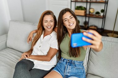 Mother and daughter smiling confident and hugging each other making selfie by the smartphone at home