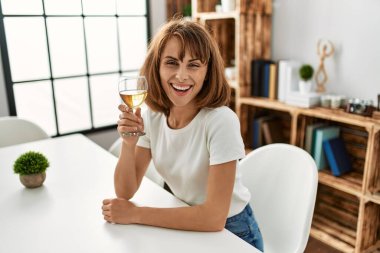 Beyaz bir kadın şarap içiyor. Evdeki masada oturuyor.