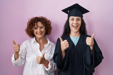 Latin kökenli anne ve kız mezuniyet şapkası ve tören cübbesi giyerek başarıya imza atıyorlar. Elleriyle olumlu jest yapıyorlar, gülümsüyorlar ve mutlular. Neşeli ifade ve kazanan jest. 