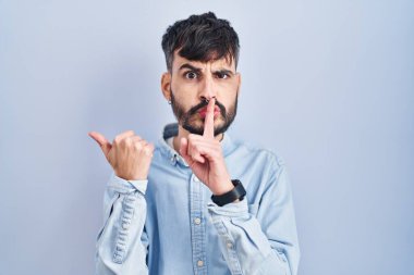 Young hispanic man with beard standing over blue background asking to be quiet with finger on lips pointing with hand to the side. silence and secret concept. 