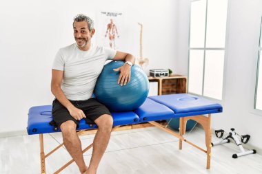 Orta yaşlı İspanyol bir adam acı dindirme kliniğinde elinde pilates topu yüzlerinde gülümseme ve doğal bir ifadeyle yan yana bakıyor. Kendinden emin gülen. 