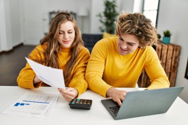 Genç beyaz çift evde dizüstü bilgisayar kullanarak aile muhasebesi yaparken gülümsüyor.