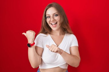 Genç beyaz kadın kırmızı arka planda dikiliyor. Arkayı işaret ediyor. Elleri havada, kendinden emin gülümsüyor. 