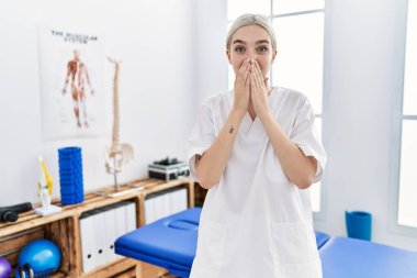 Young caucasian woman working at pain recovery clinic laughing and embarrassed giggle covering mouth with hands, gossip and scandal concept 
