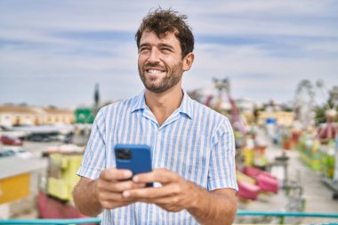 Genç İspanyol adam eğlence parkında akıllı telefon kullanarak mutlu bir şekilde gülümsüyor..