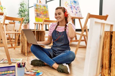 Sanat stüdyosunda dizüstü bilgisayar ve kulaklık kullanan kendine güvenen genç bir kadın.