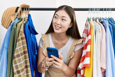 Perakende dükkanında akıllı telefon kullanan Asyalı genç bir kadın kıyafet arıyor.
