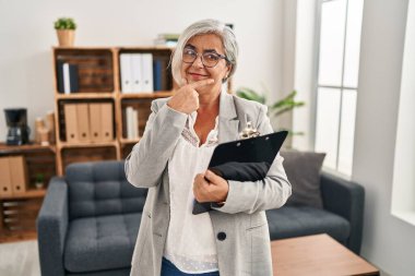 Middle age woman with grey hair at consultation office with hand on chin thinking about question, pensive expression. smiling and thoughtful face. doubt concept. 