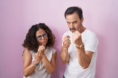 Middle age hispanic couple together over pink background ready to fight with fist defense gesture, angry and upset face, afraid of problem 