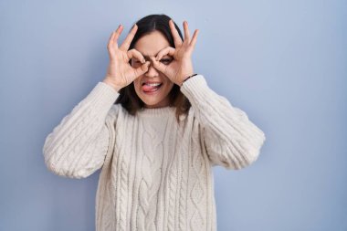 Mavi arka planda duran genç esmer kadın dürbün gibi jest yapıyor. Dürbün dilini çıkarıyor, gözler parmaklarından bakıyor. çılgın ifade. 