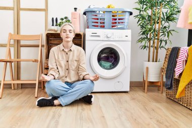 Young caucasian woman doing yoga waiting for washing machine at laundry room