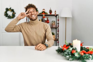 Masada sakallı genç yakışıklı adam Noel süslemeleri ile oturuyor. Barış sembolü yapıyor. Yüzünde parmaklar, gülümseyerek zaferi gösteriyor. 