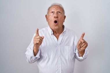 Senior man with grey hair standing over isolated background amazed and surprised looking up and pointing with fingers and raised arms. 