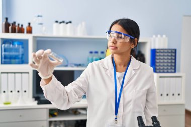 Laboratuvarda elinde bitki örneği tutan bilim adamı üniforması giyen genç Latin kadın.