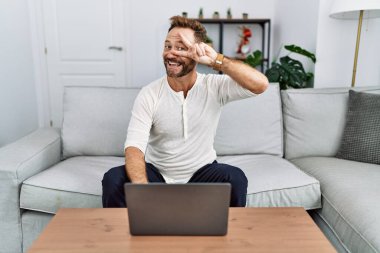 Middle age man using laptop at home doing peace symbol with fingers over face, smiling cheerful showing victory 