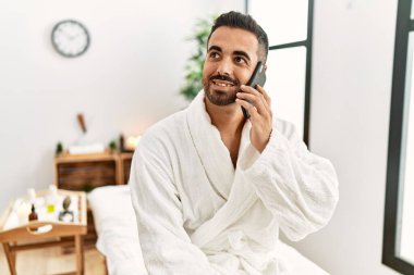 Young hispanic man wearing bathrobe talking on the smartphone at beauty center
