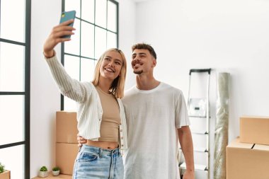 Genç beyaz çift mutlu bir şekilde gülümsüyor. Yeni evlerinde akıllı telefonların yanında selfie çekiyorlar..
