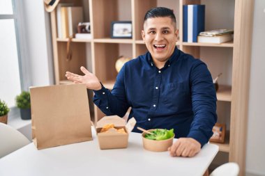 Hispanic young man eating take away food celebrating achievement with happy smile and winner expression with raised hand 