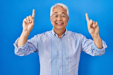 Hispanic senior man wearing glasses smiling amazed and surprised and pointing up with fingers and raised arms. 