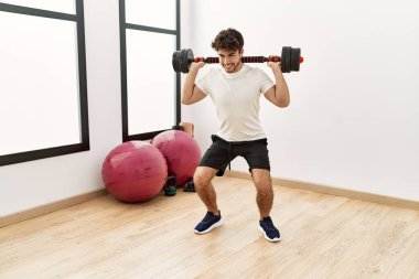 Genç İspanyol adam gülümsüyor. Spor merkezinde halterle antrenman yapıyor.