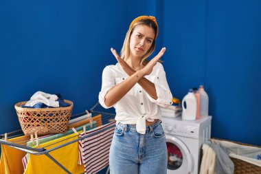 Young blonde woman at laundry room rejection expression crossing arms doing negative sign, angry face 