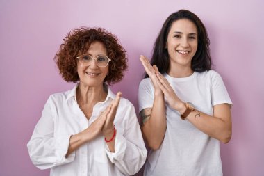 İspanyol anne ve kızı pembe arka plan üzerine gündelik beyaz tişört giyip mutlu ve neşeli, gururlu elleri alkışlıyorlar. 