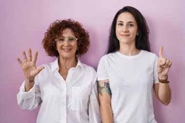 İspanyol anne ve kızı pembe arka plan üzerine gündelik beyaz tişört giyip altı numaralı parmaklarıyla işaret ederken kendinden emin ve mutlu bir şekilde gülümsüyor.. 