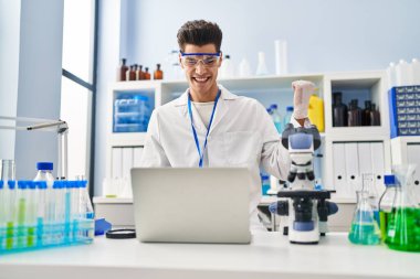 Young hispanic man working at scientist laboratory doing video call screaming proud, celebrating victory and success very excited with raised arm 