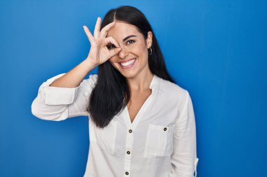 Mavi arka planda duran genç İspanyol kadın el hareketleriyle gülümsüyor ve mutlu yüzüyle parmaklarının arasından bakıyor.. 